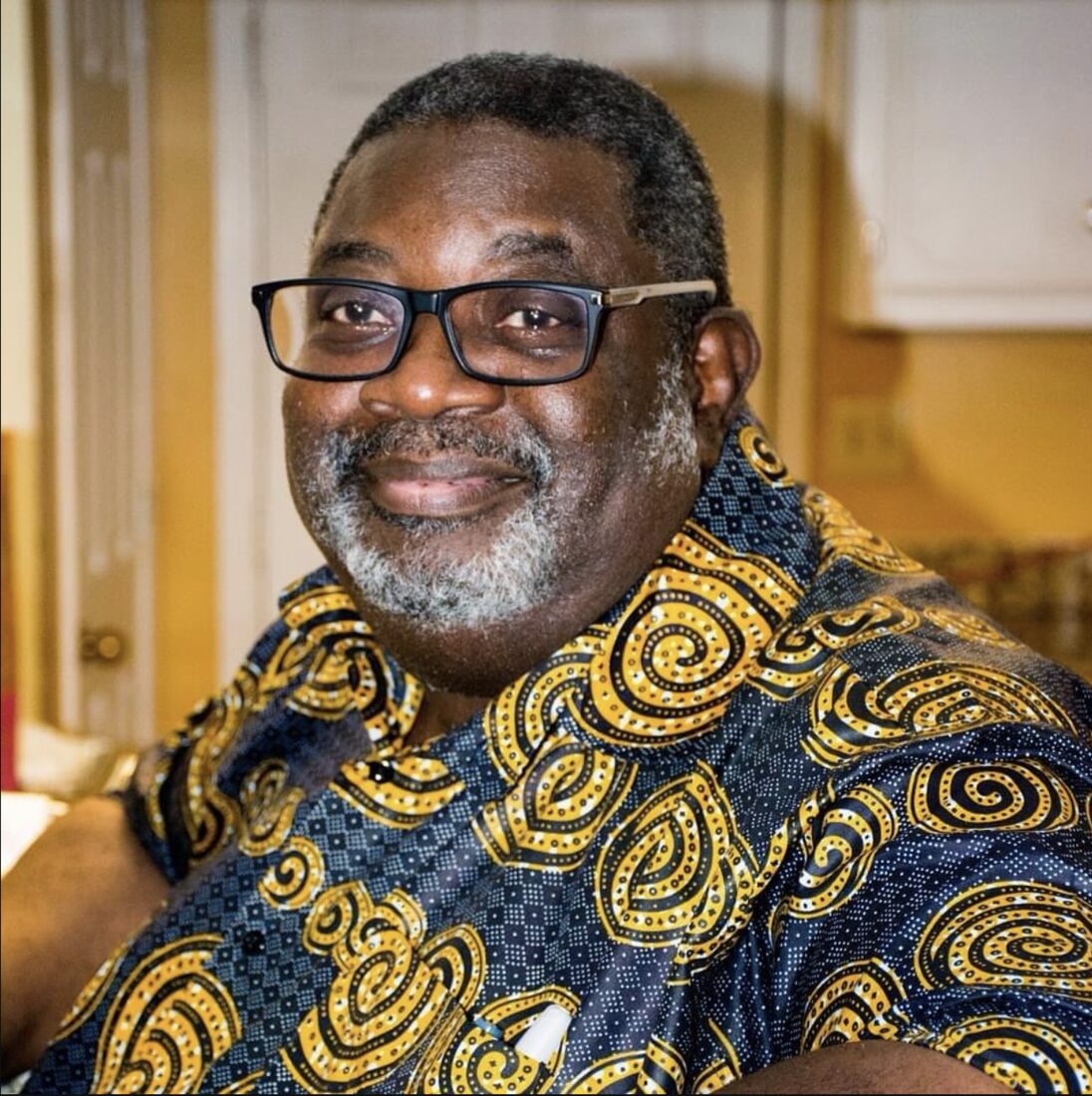 Bishop Dr. William BGK Harris smiling, wearing glasses and a navy and gold African print shirt